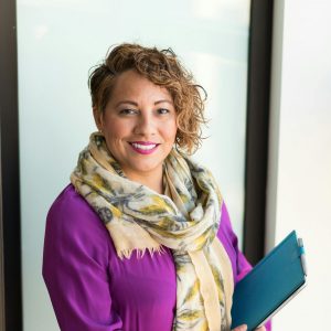 Smiling woman in a purple dress and scarf holding a tablet indoors.
